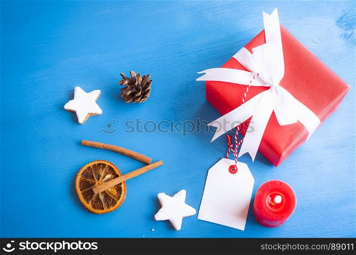 Xmas image with cinnamon sticks, star shaped cookies, dried orange, a lit candle and a lovely red gift tied with white ribbon and bow on a blue wooden table.
