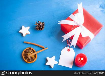 Xmas image with cinnamon sticks, star shaped cookies, dried orange, a lit candle and a lovely red gift tied with white ribbon and bow on a blue wooden table.