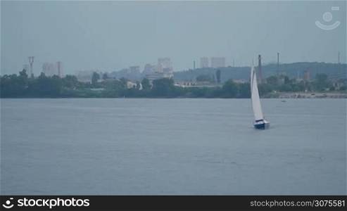 Yacht sailing on a river. The wind blows into its sails. Float boat picks up speed. Industrial landscape of big city on the background