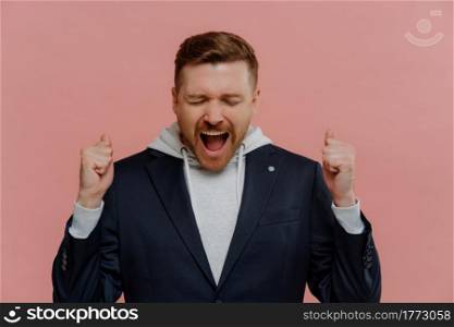 Yeah, i did it. Studio shot of overjoyed happy redhead man in casual wear keeping eyes closed from happiness and raising clenched fists while celebrating success, people reaching personal goals. Overjoyed man raising clenched fists and screaming from happiness, celebrating success