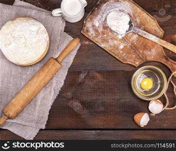 yeast dough in a wooden bowl on a brown table and ingredients, top view, empty space in the middle