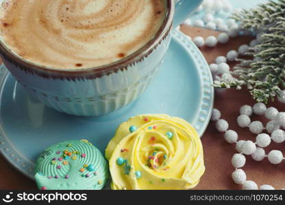 Yellow and peppermint meringues near a cup of coffee. Spruce artificial branch and Christmas beads on a craft background.