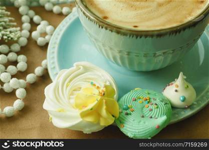 Yellow and peppermint meringues near a cup of coffee. Spruce artificial branch and Christmas beads on a craft background.