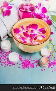 Yellow bowl with water and pink orchid flowers , white towel , lotion bottle and cream jars on mint blue wooden background. Spa, wellness or body care concept