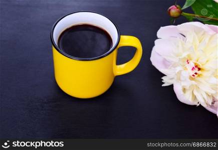 Yellow cup with black coffee on a black background, side of a white peony bud, top view