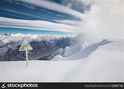 Yellow danger sign, winter mountains, blue sky and snow blizzard coming on background