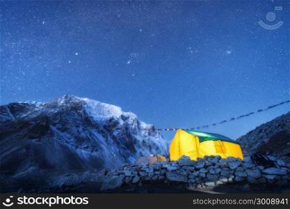 Yellow glowing tent against high rocks with snowy peak and sky with stars at night in Nepal. The Himalayan Mountains. Landscape with mountains, starry sky and bivouac. Trekking in Himalayas. Travel