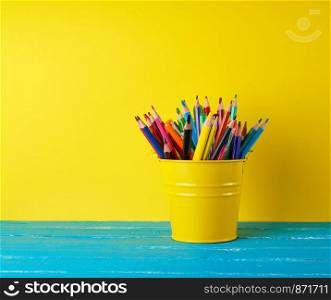 yellow green bucket with multi-colored wooden pencils and pens on a blue wooden table, concept back to school, copy space