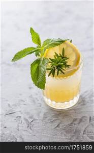 Yellow lemon shrub drink in a rocks glass garnished with mint, rosemary, and a lemon slice on a distressed and speckled light blue surface.