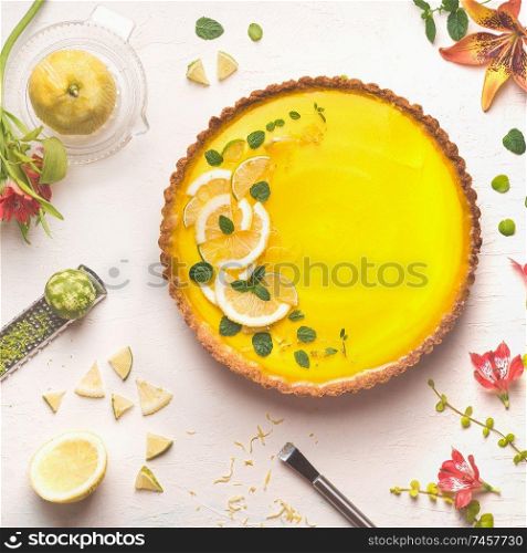 Yellow lemon tart topped with fresh lemon and lime slices on white table background with citrus ingredients and flowers, top view. Traditional french cuisine