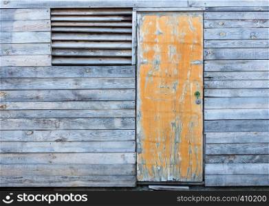 Yellow old door and a window with a wooden grate on the facade of a wooden old gray weather-beaten barn.. Wooden old weather-beaten barn with a yellow old door