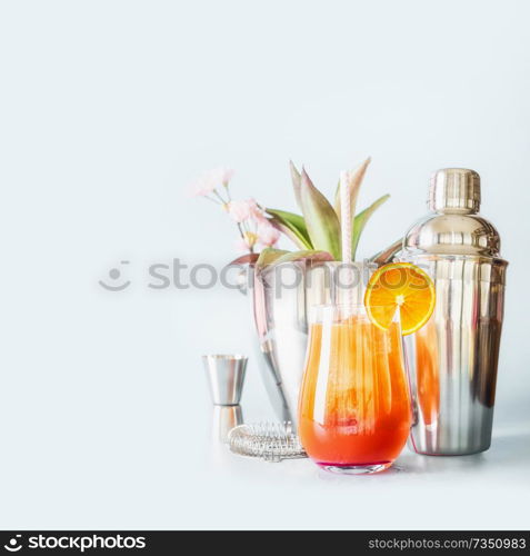 Yellow orange long drink with ice cubes, orange slice and drinking straw in glass with cocktails bar tools on table at light background with copy space.