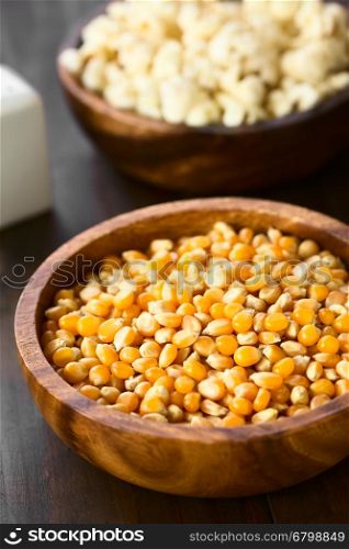 Yellow raw popcorn kernels in wooden bowl with salted popped popcorn in the back, photographed with natural light (Selective Focus, Focus in the middle of the kernels)