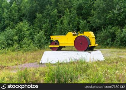 Yellow road roller for rolling asphalt on a white pedestal on a background of green bushes.