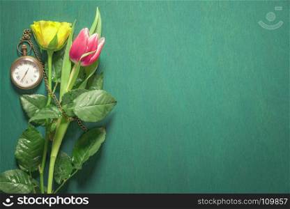 Yellow rose and a pink tulip sprinkled with water and a vintage pocket clock near them on a dark green background.