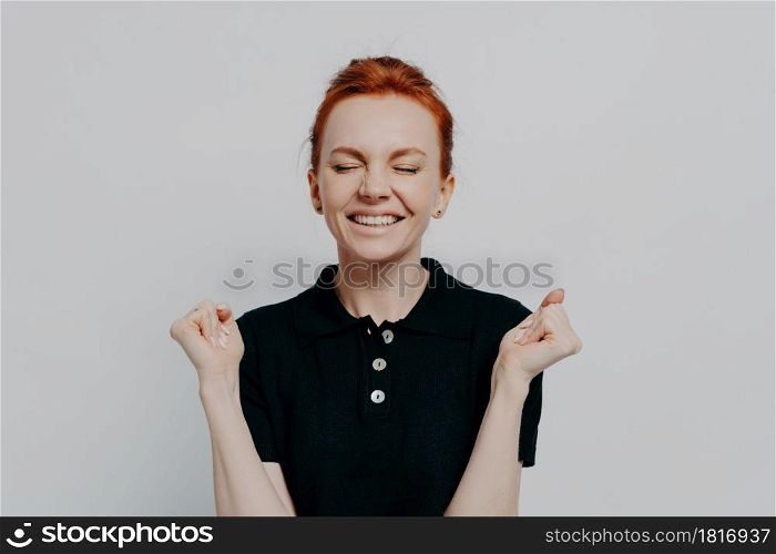 Yes, i did it. Portrait of happy euphoric red haired female with closed eyes holding clenched fists up, excited woman celebrating win, success while posing isolated over grey studio wall. Happy euphoric red haired female with closed eyes holding clenched fists up and celebrating win