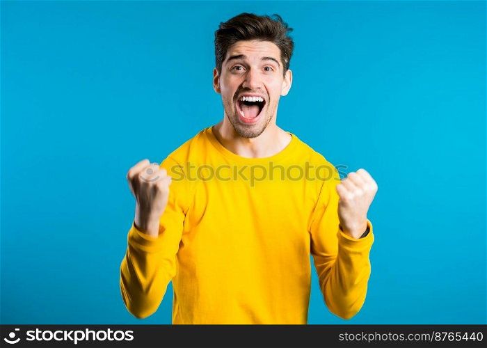 Yes winner gesture. Happy european man. Handsome guy with stylish hairdo surprised to camera over blue background.. Yes winner gesture. Happy european man. Handsome guy stylish hairdo surprised