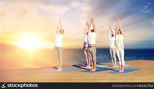 yoga and healthy lifestyle concept - group of people doing upward salute pose on wooden pier over sea background. group of people doing yoga outdoors