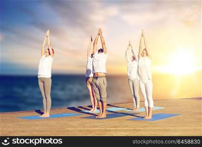 yoga and healthy lifestyle concept - group of people doing upward salute pose on wooden pier over sea background. group of people doing yoga outdoors