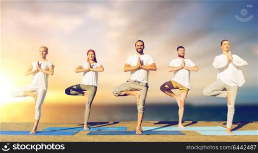yoga, fitness and healthy lifestyle concept - group of people doing tree pose on mat outdoors on wooden pier over sea background. people doing yoga tree pose on mat outdoors