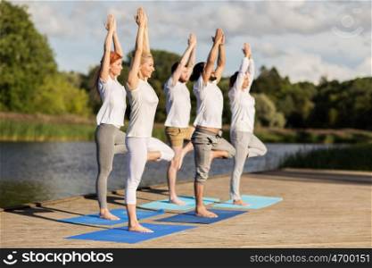 yoga, fitness, sport, and healthy lifestyle concept - group of people in tree pose on mat outdoors on river or lake berth