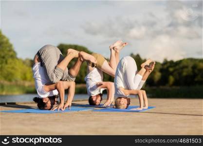 yoga, fitness, sport, and healthy lifestyle concept - group of people making tripod egg pose n mat outdoors on river or lake berth