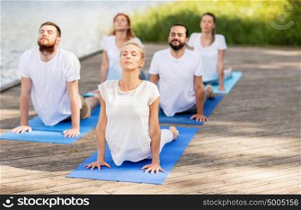 yoga, fitness, sport, and healthy lifestyle concept - group of people making upward facing dog or cobra pose on river or lake berth. group of people making yoga exercises outdoors