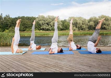 yoga, fitness, sport, and healthy lifestyle concept - group of people making supported shoulderstand pose on mat outdoors on river or lake berth. people making yoga in shoulderstand pose on mat