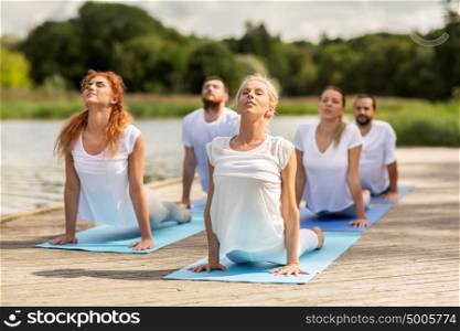 yoga, fitness, sport, and healthy lifestyle concept - group of people making upward facing dog or cobra pose on river or lake berth. group of people making yoga exercises outdoors