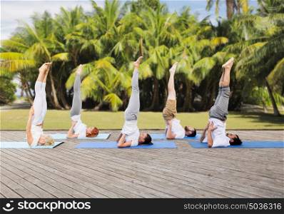yoga, fitness, sport, and healthy lifestyle concept - group of people making supported shoulderstand pose on mat outdoors over exotic natural background with palm trees. people making yoga in shoulderstand pose on mat