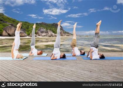 yoga, fitness, sport, and healthy lifestyle concept - group of people making supported shoulderstand pose on mat outdoors over exotic tropical beach background. people making yoga in shoulderstand pose on mat