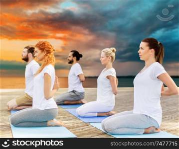 yoga, fitness, sport, and healthy lifestyle concept - group of people making hero pose on mat outdoors on sea pier over sunset background. people making yoga in hero pose outdoors
