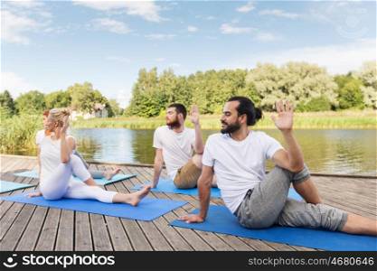 yoga, fitness, sport, and healthy lifestyle concept - group of people sitting in half lord of the fishes pose on mat outdoors on river or lake berth