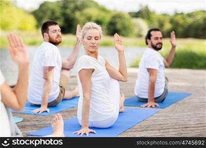 yoga, fitness, sport, and healthy lifestyle concept - group of people sitting in half lord of the fishes pose on mat outdoors on river or lake berth
