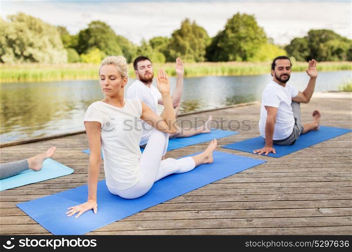 yoga, fitness, sport, and healthy lifestyle concept - group of people sitting in half lord of the fishes pose on mat outdoors on river or lake berth. people making yoga in half lord of the fishes pose