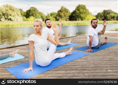 yoga, fitness, sport, and healthy lifestyle concept - group of people sitting in half lord of the fishes pose on mat outdoors on river or lake berth. people making yoga in half lord of the fishes pose