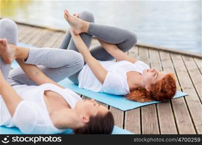 yoga, fitness, sport, and healthy lifestyle concept - women making supine pigeon pose on mat outdoors on river or lake berth