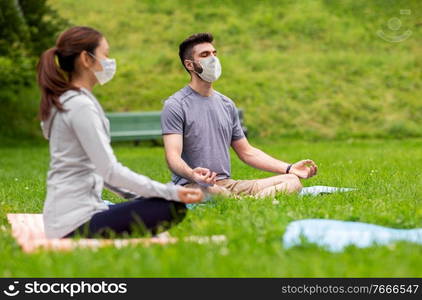 yoga, health and pandemic concept - people wearing face protective medical mask for protection from virus disease meditating in lotus pose at summer park. people in masks meditating at summer park