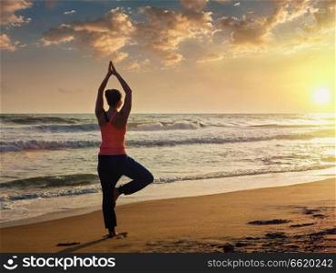 Yoga outdoors - sporty fit woman doing Hatha yoga asana Vrikshasana tre pose on tropical beach on sunset. Young sporty fit woman doing yoga tree asana on beach
