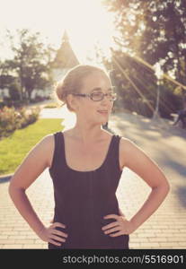 young 20s blond haired women posing with hands on her waist outdoors weared black tank top in glasses. Shi is very pretty and fresh looking. Backlit