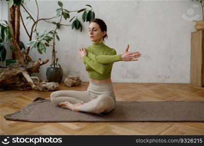 Young active woman exercising while sitting on yoga mat stretching arms in living room at home. Young woman exercising while sitting on yoga mat stretching arms
