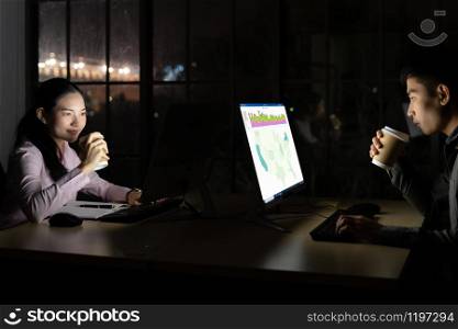 Young adult asian businessman and woman drinking hot cofee while working late at night in their office with desktop computer and laptop. Using as hard working and working late concept.