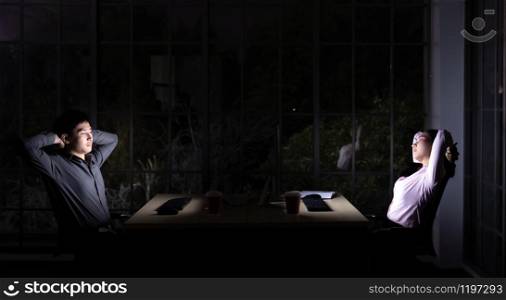 Young adult asian businessman and woman stretching while working late at night in their office with desktop computer and laptop. Using as hard working and working late concept.