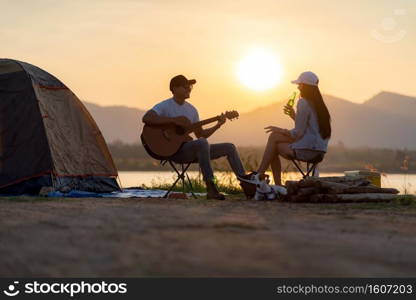 Young adult Asian couple playing guitar and drinking beer beside their tent c&site while sunset. C&ing trip with dog outdoor activity c&site concept.