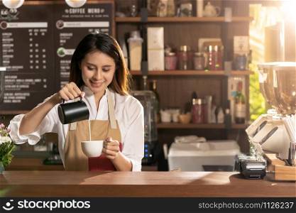 Young adult asian female barista pouring fresh milk to prepare latte coffee for customer in cafe bar.