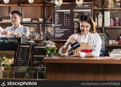 Young adult asian female barista pouring fresh milk to prepare latte coffee for customer in cafe bar.