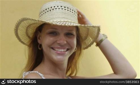 Young adult blonde woman in straw hat laughing against yellow wall