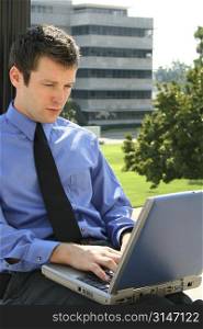 Young adult business man using a laptop computer with an office building in the background.