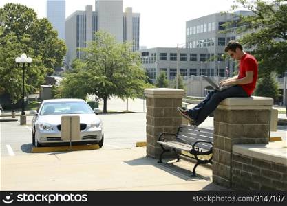 Young adult casually dressed male sitting on a wall with a laptop computer.