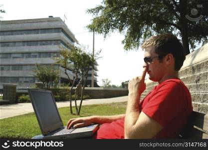 Young adult casually dressed male using a laptop computer on a park bench with his finger in his nose.